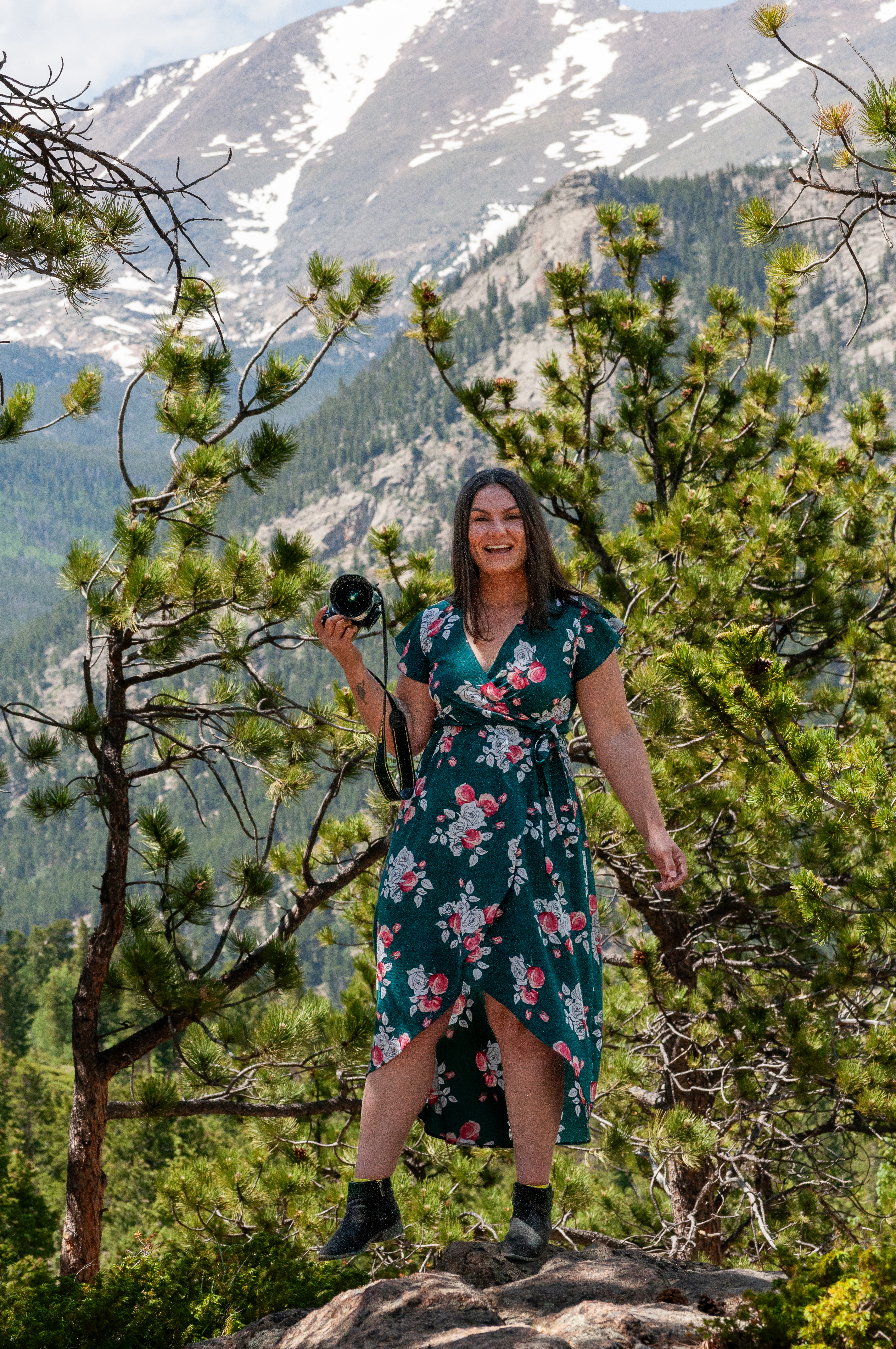 portrait of the photographer, she is smiling and looking at the camera while holding a camera near her right shoulder. trees and mountains are in the background.